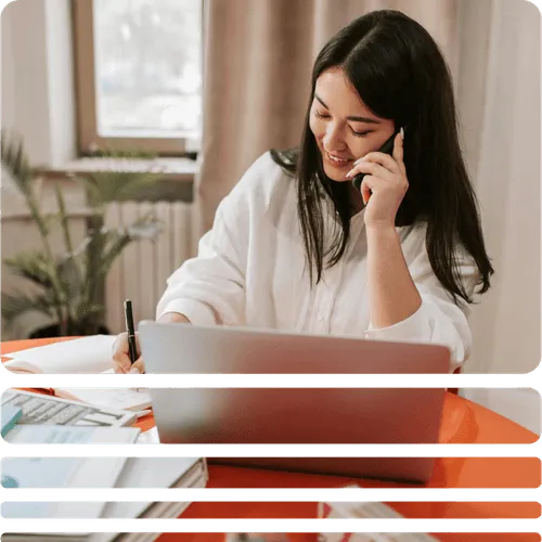 Smiling woman with long dark hair talking on a phone while writing notes at a desk with a laptop and books.