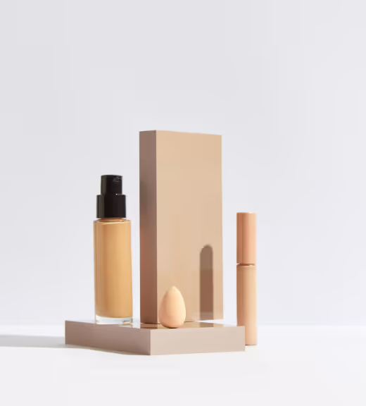 Minimalist makeup set including a liquid foundation bottle with a pump, a teardrop makeup sponge, and a tube, arranged on and beside beige geometric blocks.