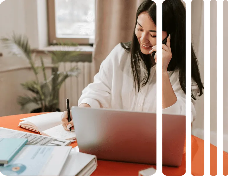 Smiling woman talking on phone while writing notes and working on laptop at a desk with documents.