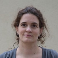 Portrait d'une femme aux cheveux bruns attachés, portant un haut gris, avec un léger sourire et un fond beige flou.