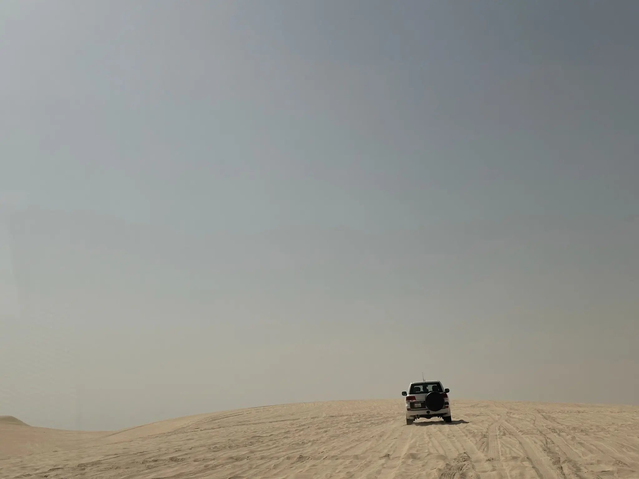White SUV driving over sandy desert dunes under a clear sky.