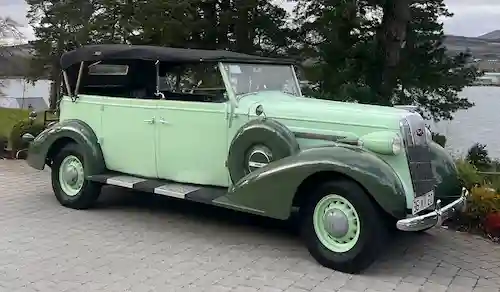 Vintage mint green convertible car with black soft top parked on a paved area near trees and water.