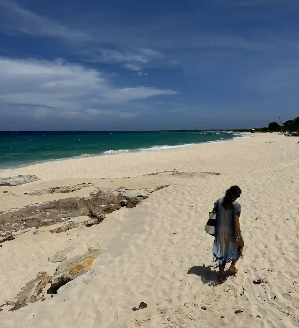 Barrett walking barefoot on a sandy beach near turquoise ocean under a blue sky with scattered clouds.