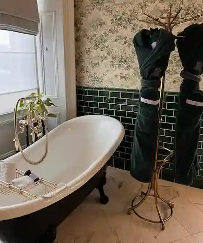 Elegant bathroom with a black clawfoot bathtub, gold fixtures, floral wallpaper, dark green tiles, and two dark green robes on a gold stand.