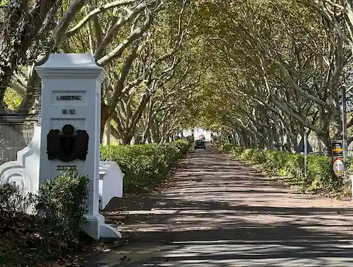 Tree-lined driveway with dense green foliage forming an arch over the road and a white stone pillar marked 'Lanzherac 1692' on the left.