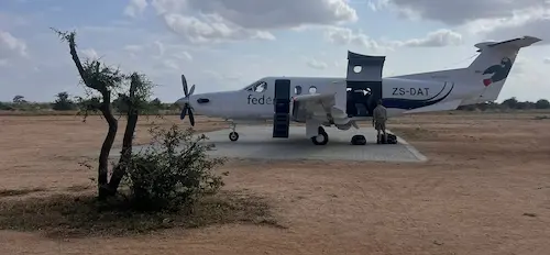 Small white propeller airplane parked on dirt with open doors and a person loading luggage under a partly cloudy sky.