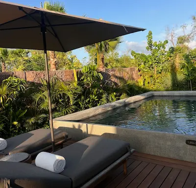 Outdoor pool area with two lounge chairs topped with rolled white towels under a large umbrella, surrounded by tropical plants.