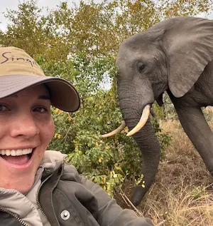 Barrett smiling wearing a hat taking a selfie near a young elephant in a natural outdoor setting.