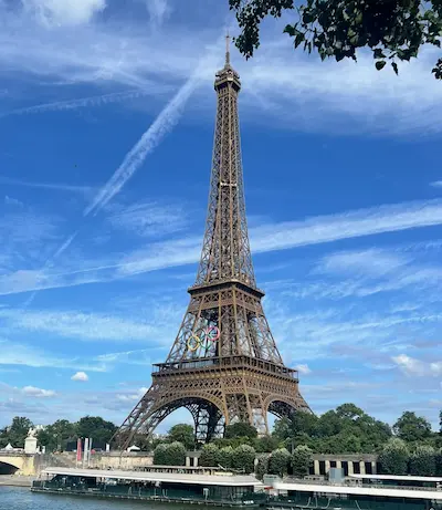 Eiffel Tower in Paris with Olympic rings displayed, under a blue sky with wispy clouds and surrounding greenery.