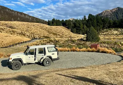 White off-road vehicle parked on a gravel area with dry grassy hills and forested mountains in the background under a blue sky with scattered clouds.