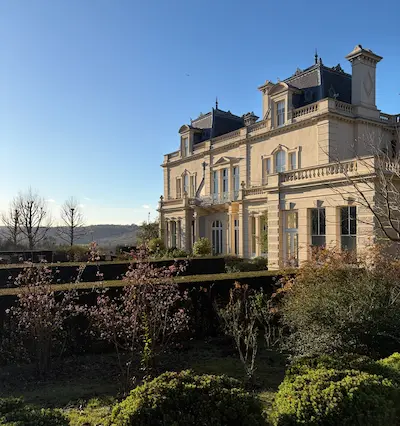 Elegant beige mansion with blue shutters surrounded by trimmed bushes and trees under a clear blue sky.