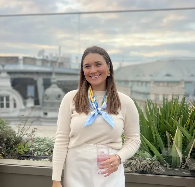 Barrett with long brown hair wearing a cream sweater and a blue patterned scarf, smiling and holding a glass with a pink drink, standing outdoors with urban buildings and plants in the background.