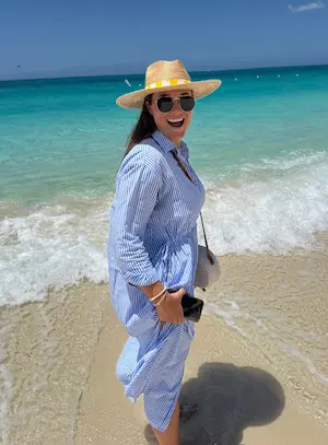 Barrett in a striped blue dress, sunglasses, and straw hat smiling on a sandy beach with turquoise sea waves behind her.