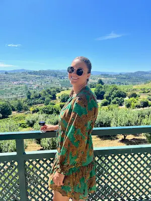 Barrett in sunglasses and green patterned dress holding a glass of red wine, standing on a balcony overlooking a sunny landscape of trees and hills under a clear blue sky.