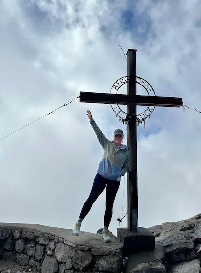 Barrett standing on rocky ledge next to a large wooden cross with cloudy sky in the background.
