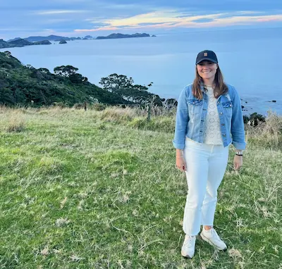 Barrett in white pants and denim jacket standing on grassy hill with ocean and islands in the background at sunset.