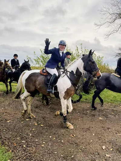 Barrett in navy jacket and helmet waving while riding a black and white horse on a dirt path with other riders nearby.