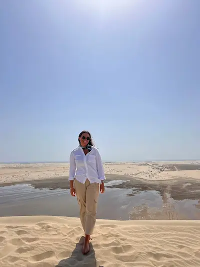 Barrett in a white shirt and beige pants standing barefoot on sand dunes under a clear blue sky with a shallow water body in the background.