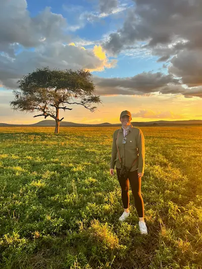 Barrett standing on grassy field at sunset with a single large tree and hills in the background under a partly cloudy sky.