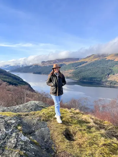 Barrett wearing a brown hat and dark jacket standing on a mossy rock overlooking a scenic lake with forested hills under a blue sky.