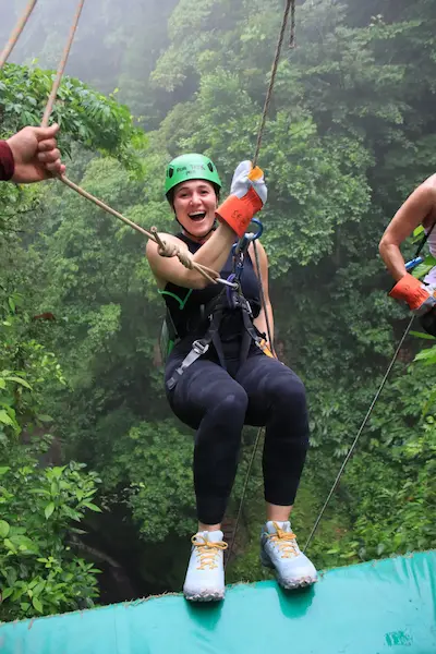 Barrett wearing a green helmet and orange gloves smiling while harnessed for a zipline over lush green forest.
