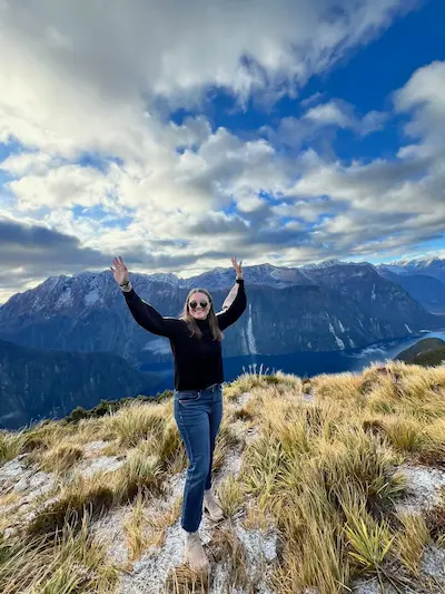 Barrett wearing sunglasses and a black sweater standing with arms raised on a grassy mountain ridge overlooking a lake and snow-capped mountains under a partly cloudy blue sky.