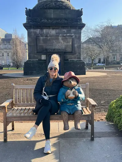 Barrett in winter clothes sitting on a bench next to a Paddington Bear statue holding a sandwich.