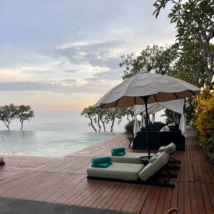 Infinity pool at sunset with lounge chairs, umbrellas, and a wooden deck overlooking the ocean and trees.