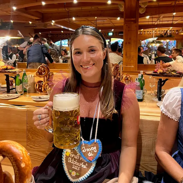 Barrett smiling in traditional Bavarian dress holding a large beer mug at an Oktoberfest celebration inside a wooden hall.
