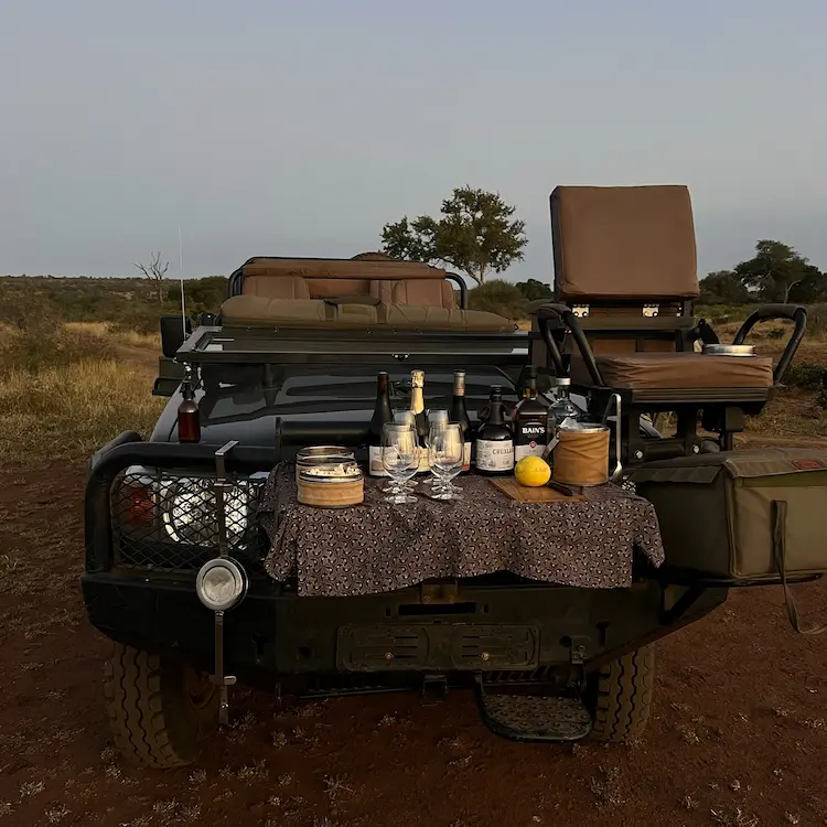 Safari vehicle with a rear table set up with bottles, glasses, snacks, and a lemon in a savannah landscape.