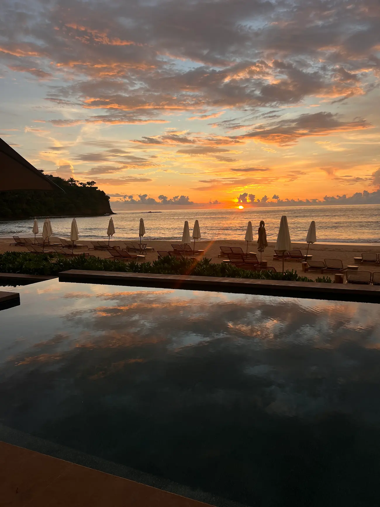 Sunset over a beach with closed umbrellas and lounge chairs, reflecting orange and blue clouds in a calm pool in the foreground.