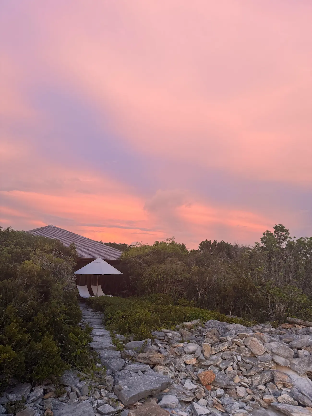 Stone pathway leading to a shaded seating area with two lounge chairs and an umbrella, surrounded by greenery under a pink and purple sunset sky.