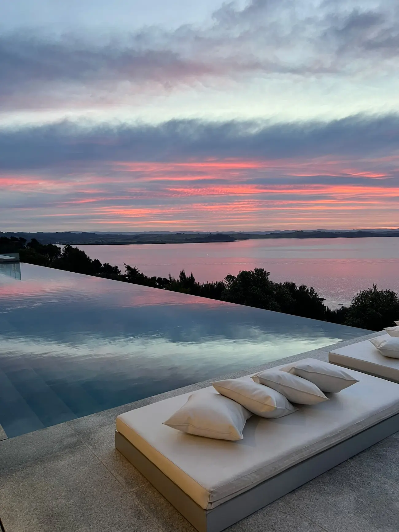 Infinity pool at sunset reflecting colorful pink and blue sky, with cushioned loungers in the foreground and a lake surrounded by hills in the background.