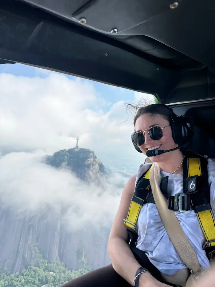 Barrett in sunglasses and headset wearing a yellow harness inside a helicopter with the Christ the Redeemer statue visible through the window amid clouds.