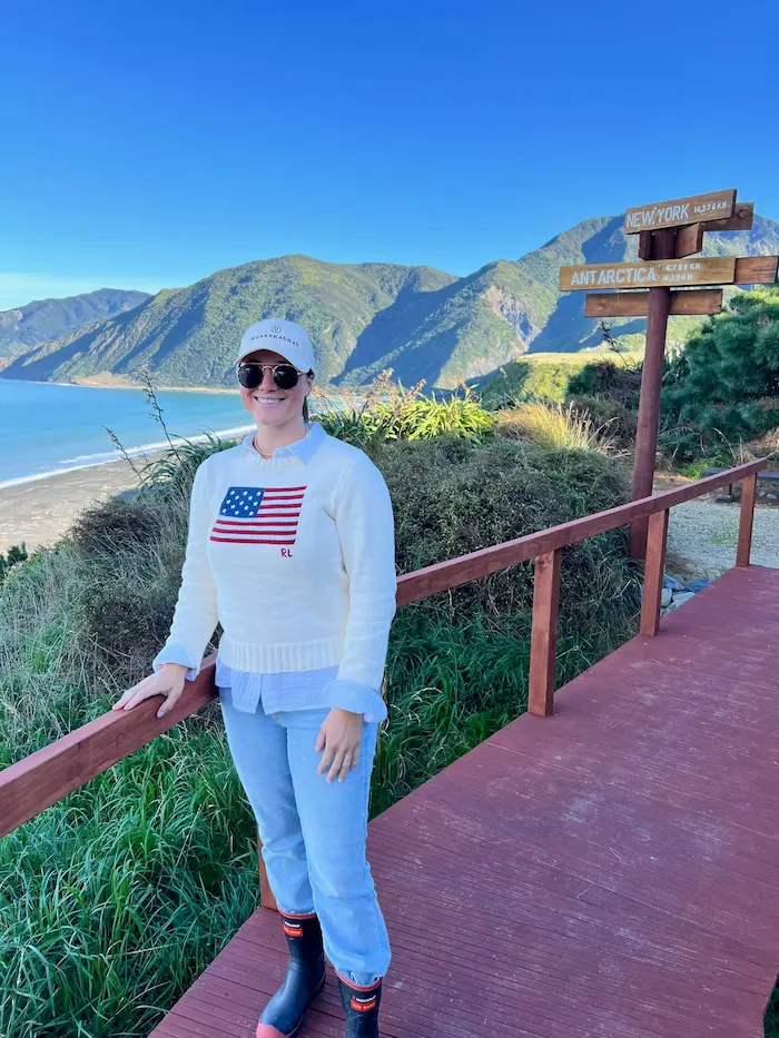 Barrett wearing sunglasses, a white sweater with an American flag, and a cap standing on a wooden path with mountains, ocean, and directional signs in the background.