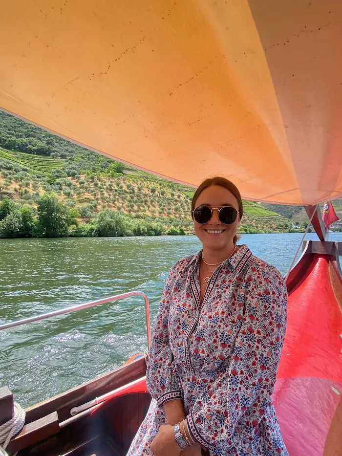 Barrett wearing sunglasses and a floral dress sitting on a red boat with a river and hillside vineyards in the background.