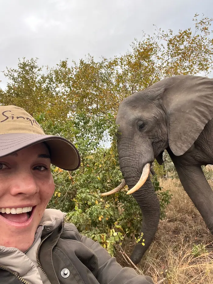 Barrett smiling wearing a hat taking a selfie with a young elephant standing next to green bushes.