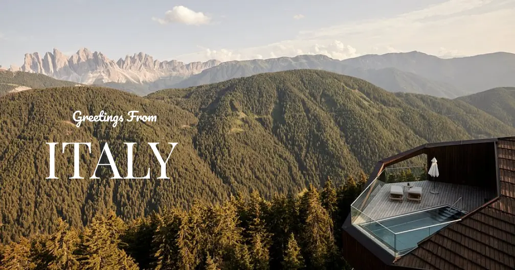 View of forested mountains in Italy with a modern balcony pool and two lounge chairs on the right.