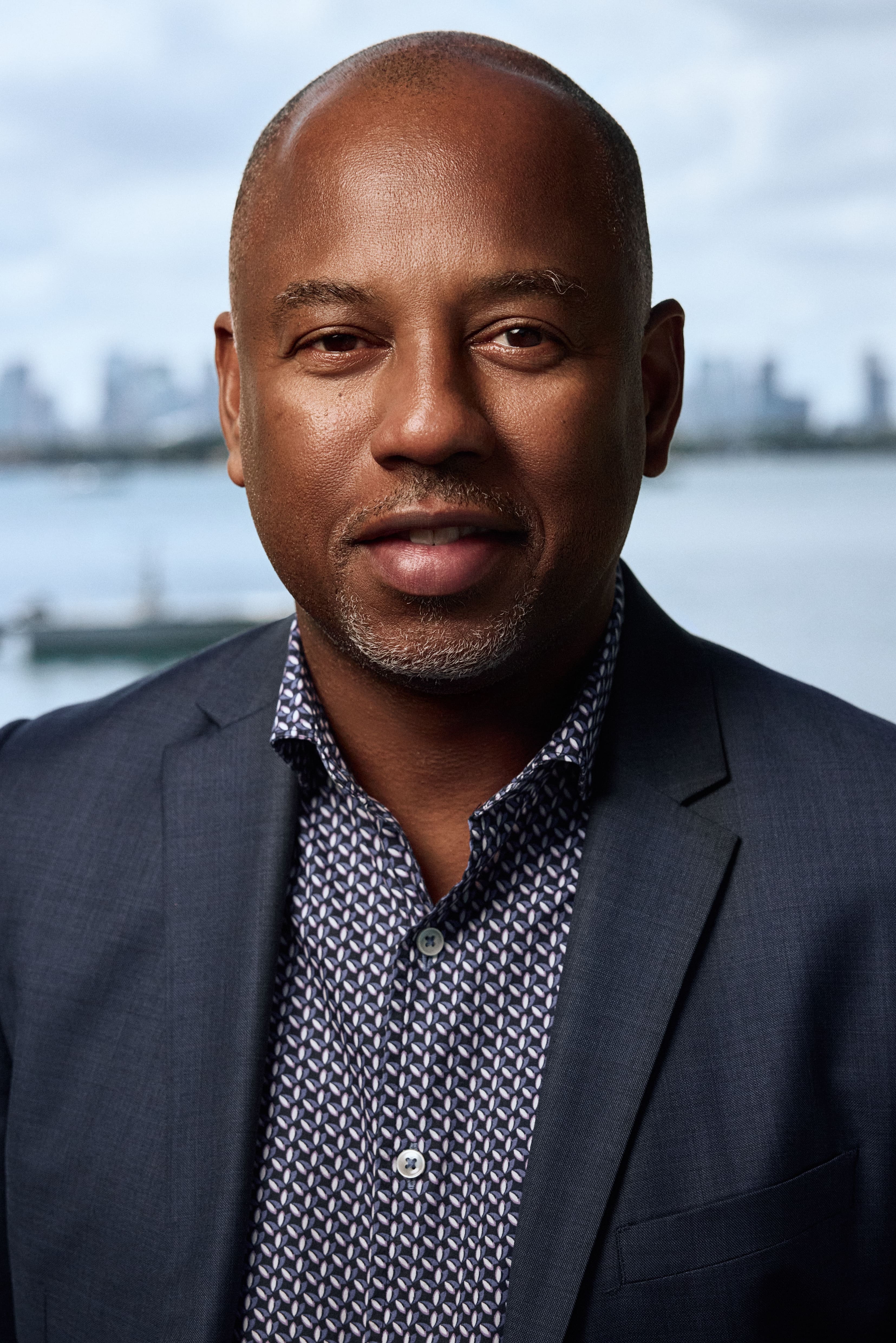 Close-up of a smiling man wearing a patterned shirt and dark blazer with a blurred waterfront cityscape in the background.