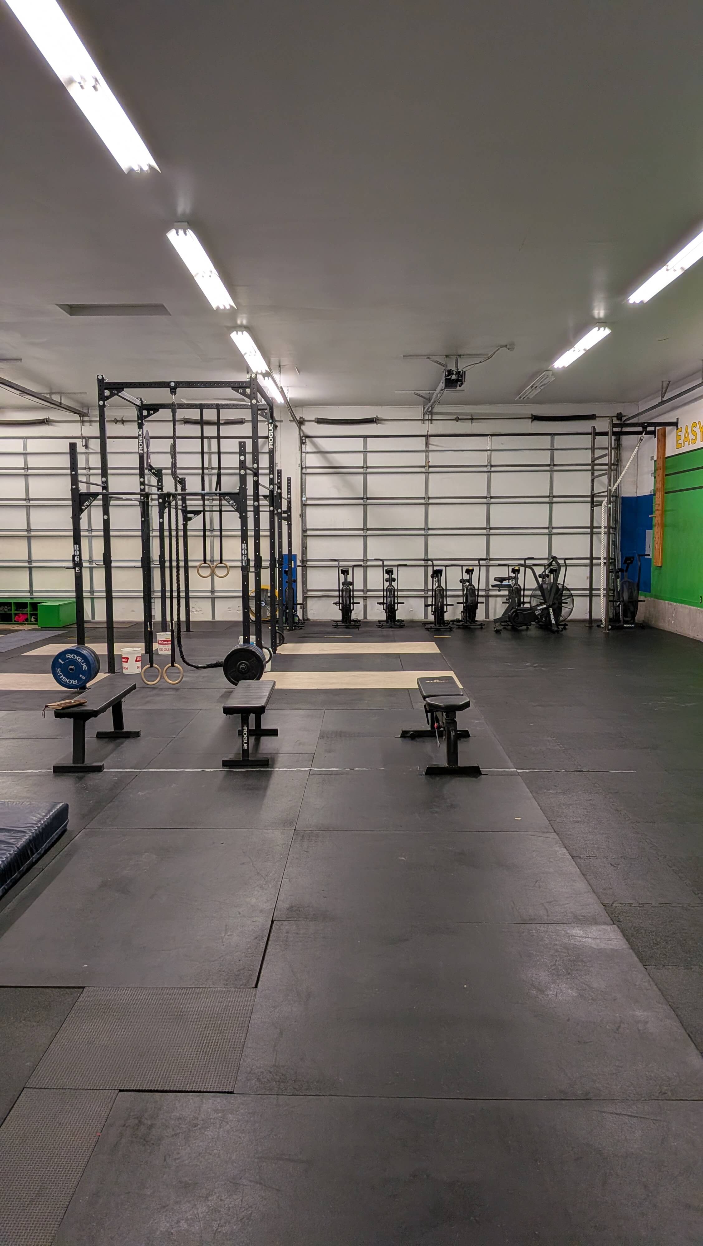 Empty gym with black rubber flooring, workout benches, a pull-up rig with rings, rowing machines, and a green wall on the right side. Trailhead Physical Therapy and Performance