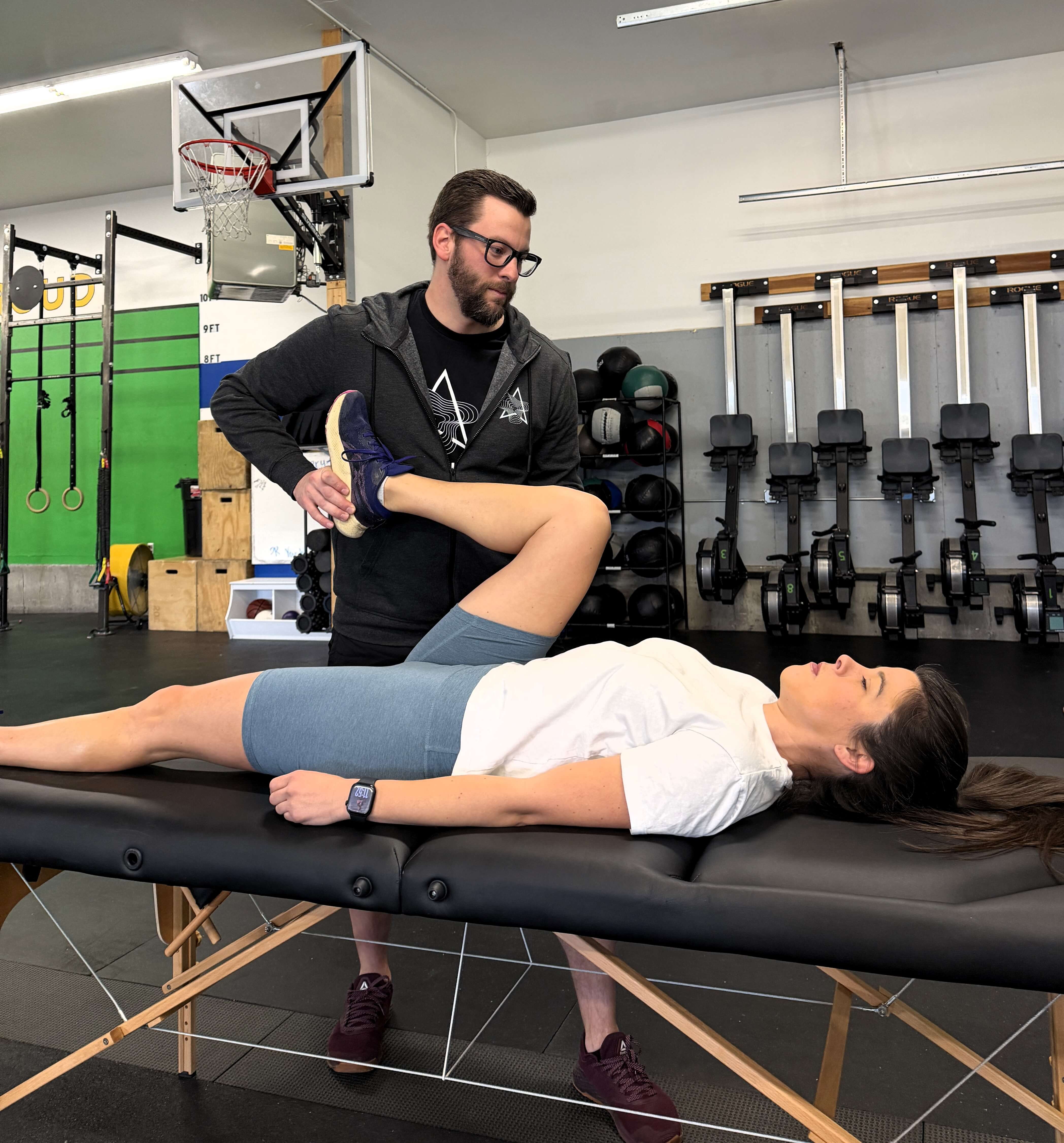 Physical therapist assisting woman lying on a massage table with bent knee stretch in a gym setting. trailhead-physical-therapy