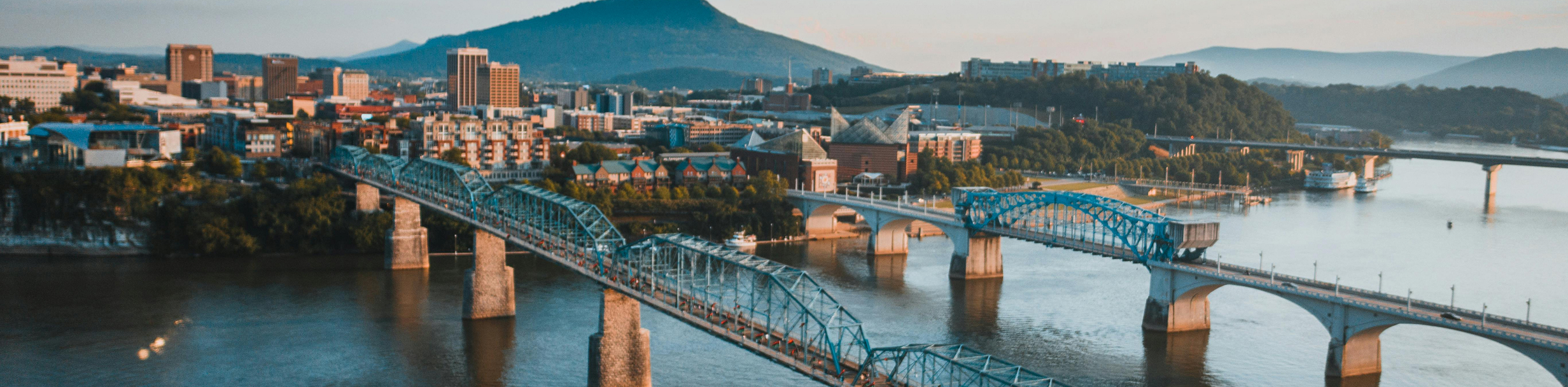 Aerial view of a Chattanooga with multiple bridges crossing a river, mountains in the background, and buildings lining the waterfront.