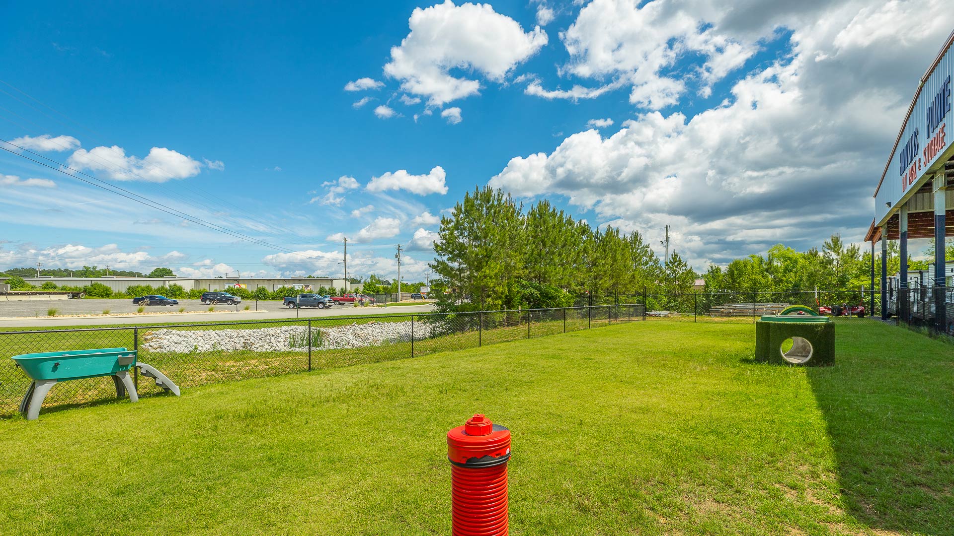 Outdoor fenced dog park with green grass, a red fire hydrant, a green tunnel, and a bath station.