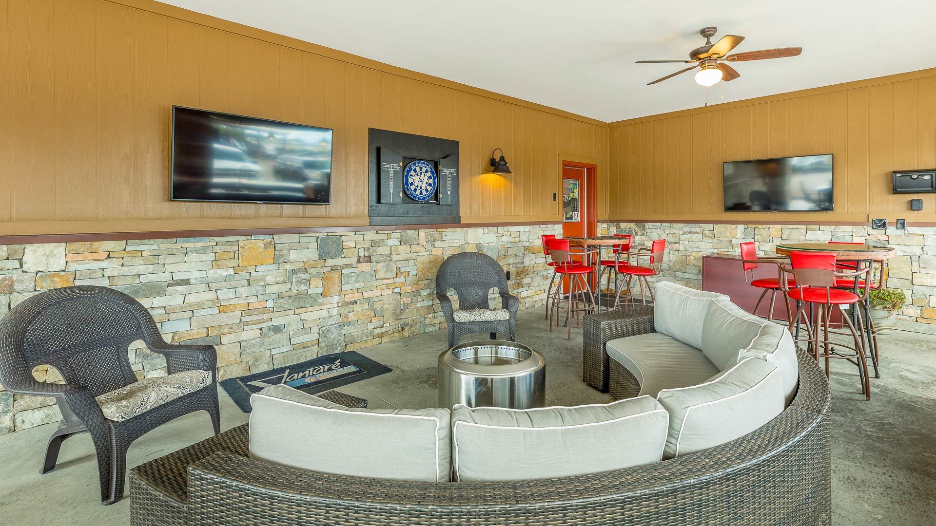 Covered outdoor seating area with curved cushioned wicker sofa, black chairs, two wall-mounted TVs, a dartboard, and high tables with red chairs.