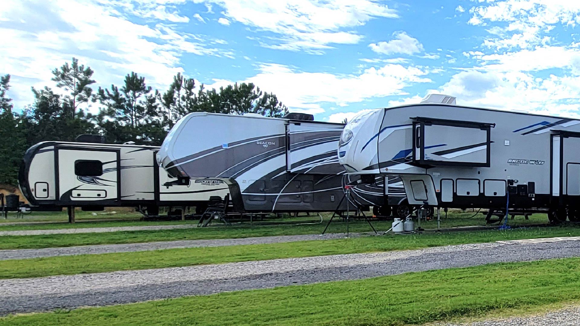 Three parked fifth-wheel long term RV trailers on grassy lots under a partly cloudy sky.