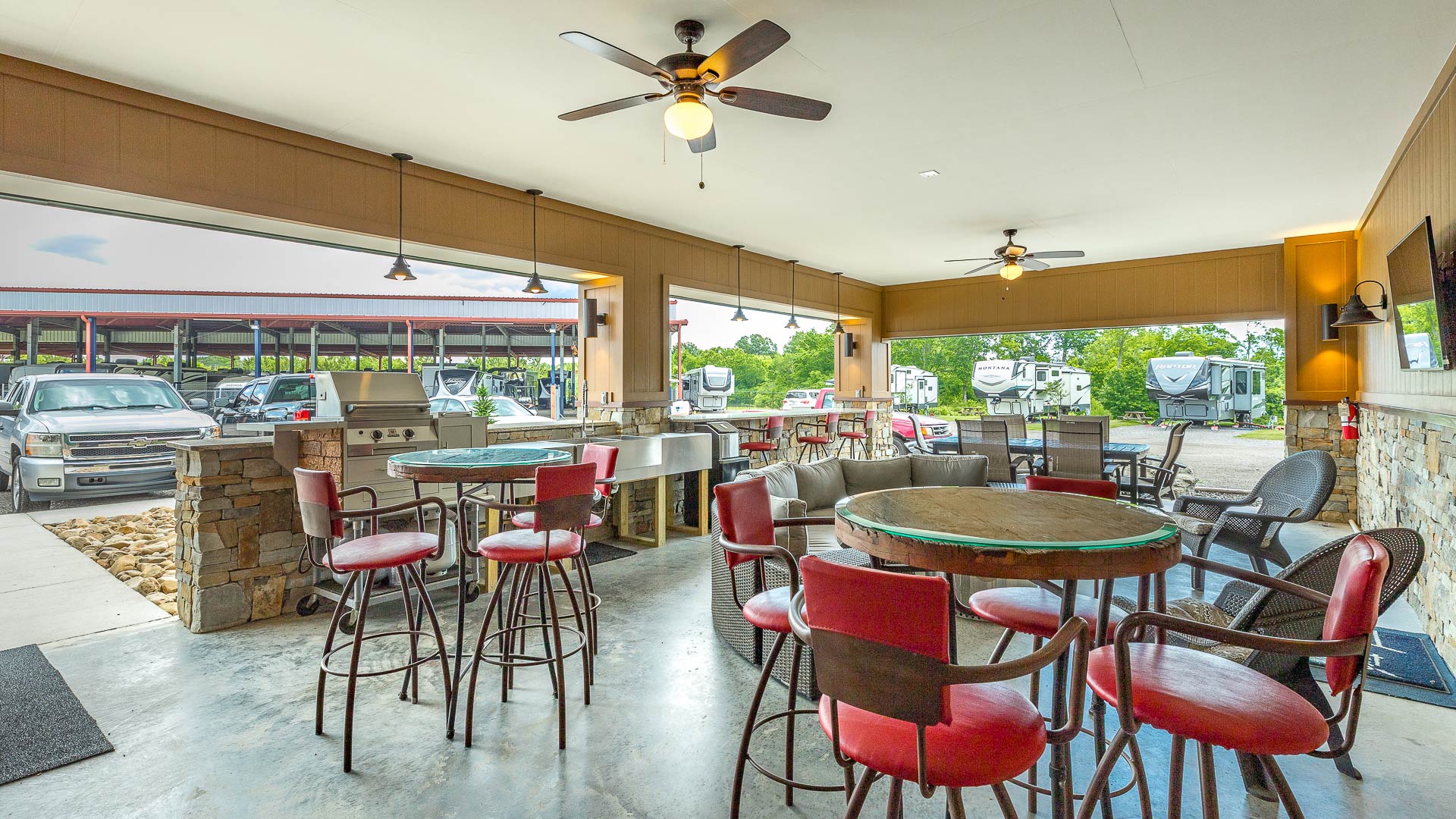 Covered outdoor seating area with round tables, red cushioned bar stools, ceiling fans, and a grill, overlooking parked trucks and RVs.