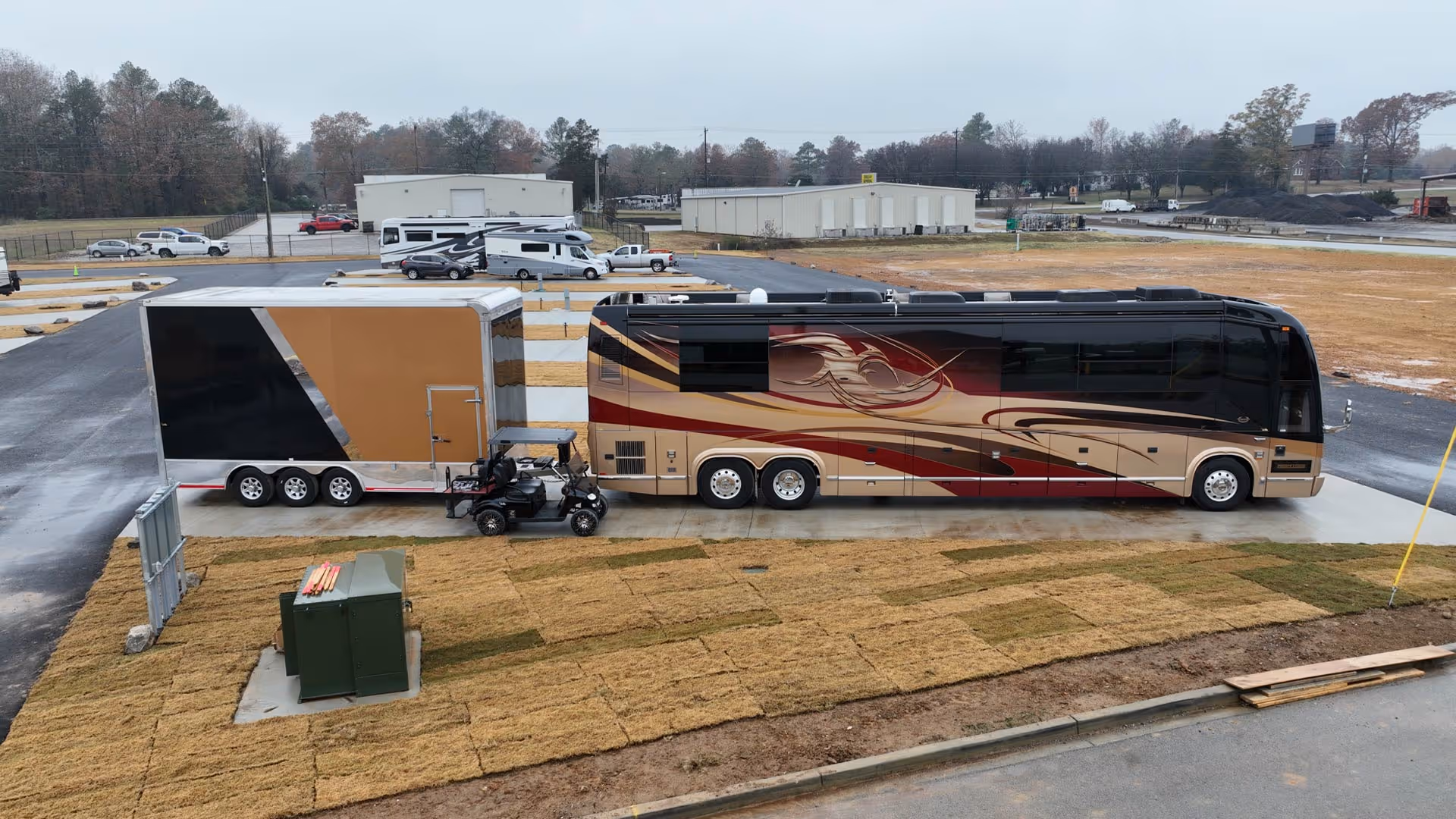 Large beige and maroon motorhome with a decorative design parked next to a three-axle trailer and a black golf cart on a concrete pad.