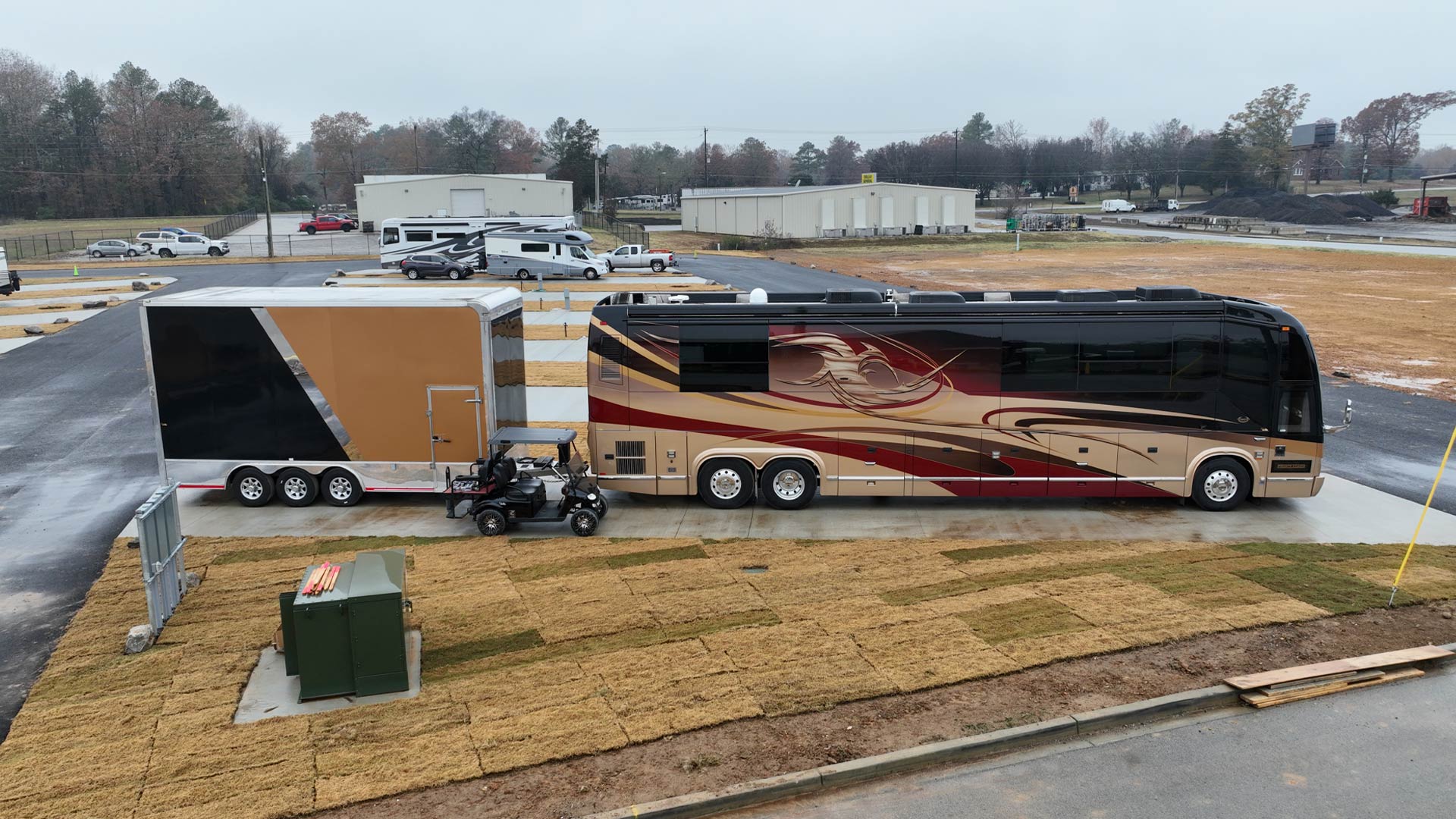 Large beige and maroon motorhome with a decorative design parked next to a three-axle trailer and a black golf cart on a concrete pad.