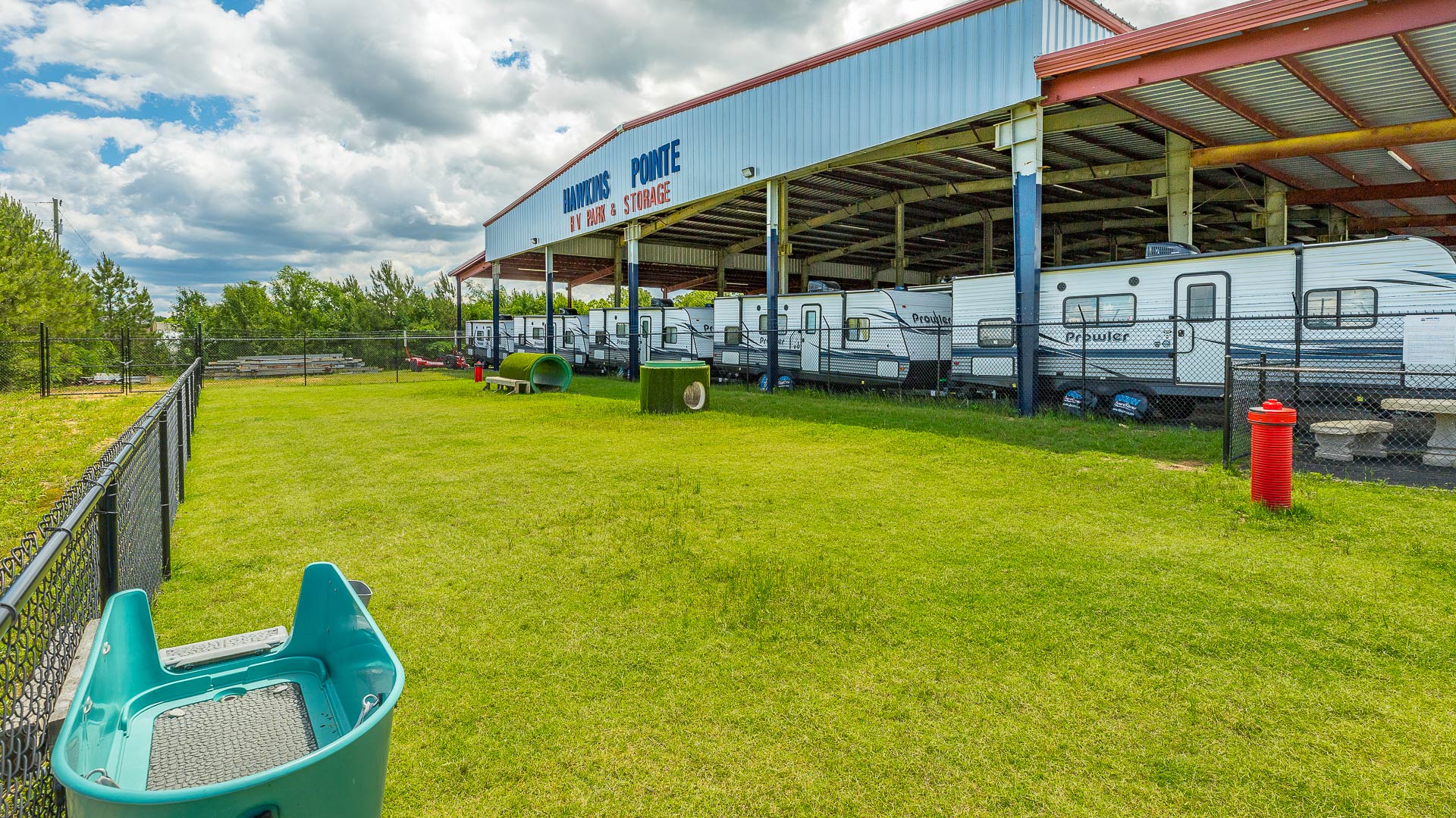Fenced green grassy dog park with agility equipment next to a covered storage area for RVs.