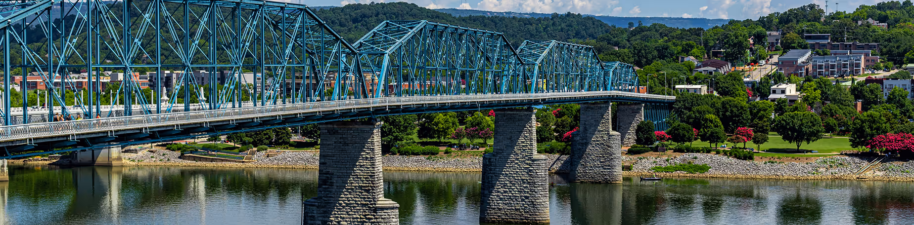 Chattanooga bridge crossing the river with green trees and buildings in the background under a blue sky.